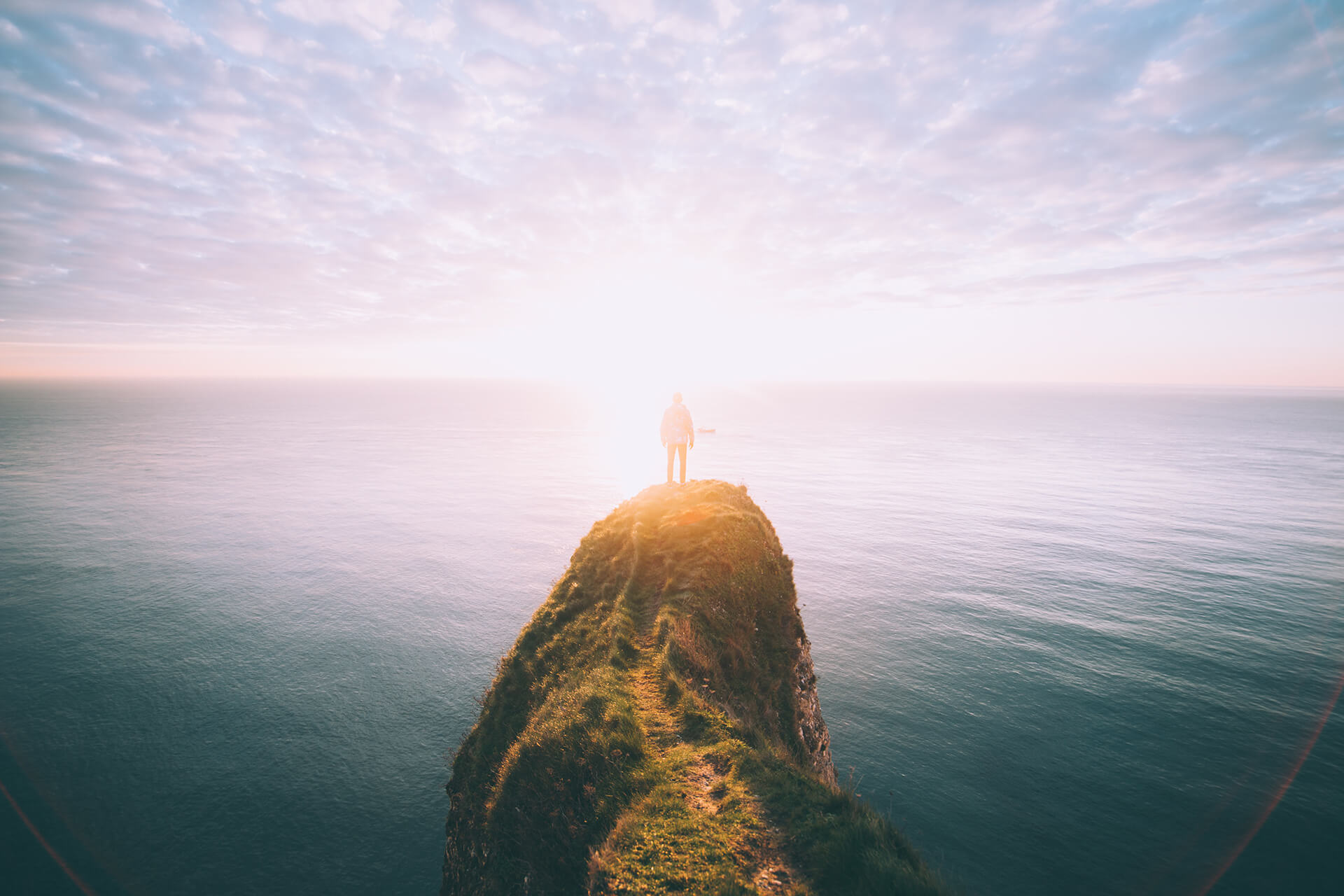 A person standing on the tip of a grassy headland, looking out over a calm sea at sunrise
