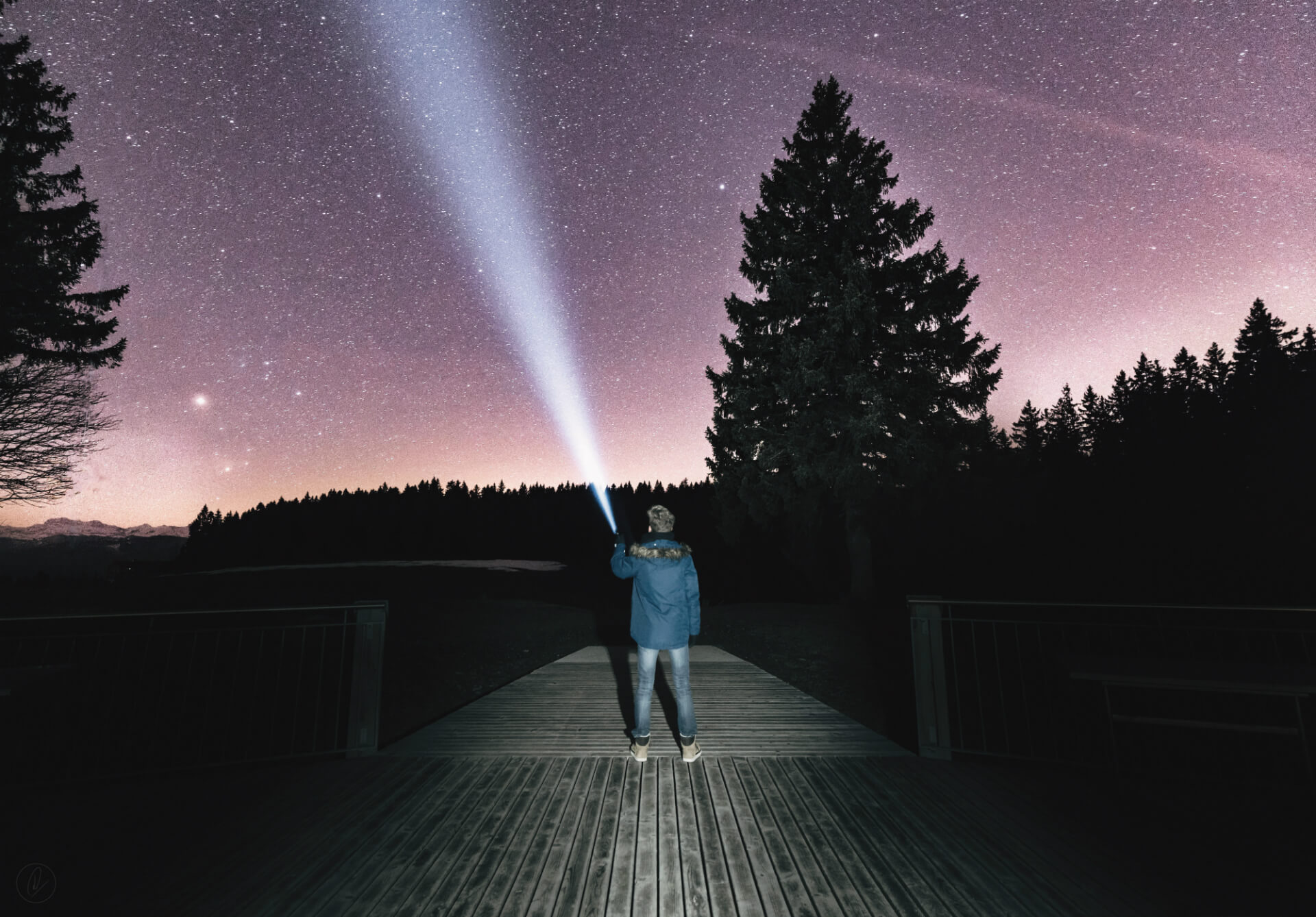 A person on a wooden boardwalk at night, shining a torch beam up into a starry sky