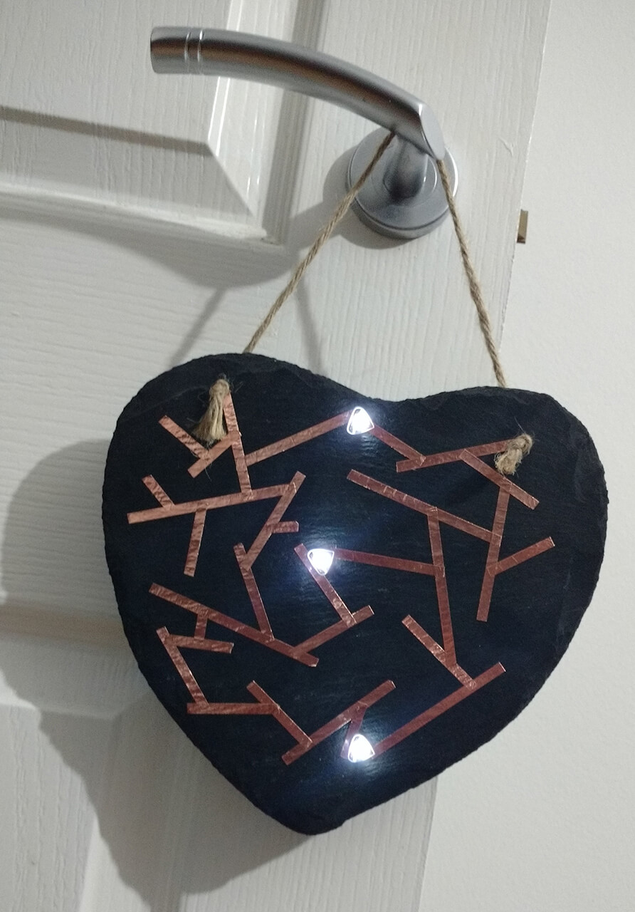 A heart-shaped slate hanging from a door handle, decorated with a copper tape pattern and three lit LED stickers