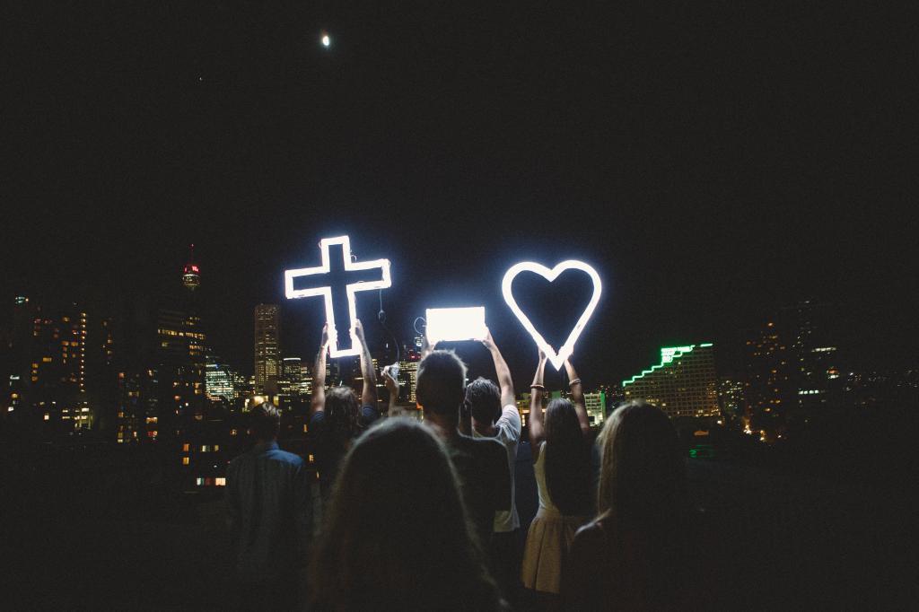 A crowd at night holding up glowing neon signs shaped like a cross and a heart