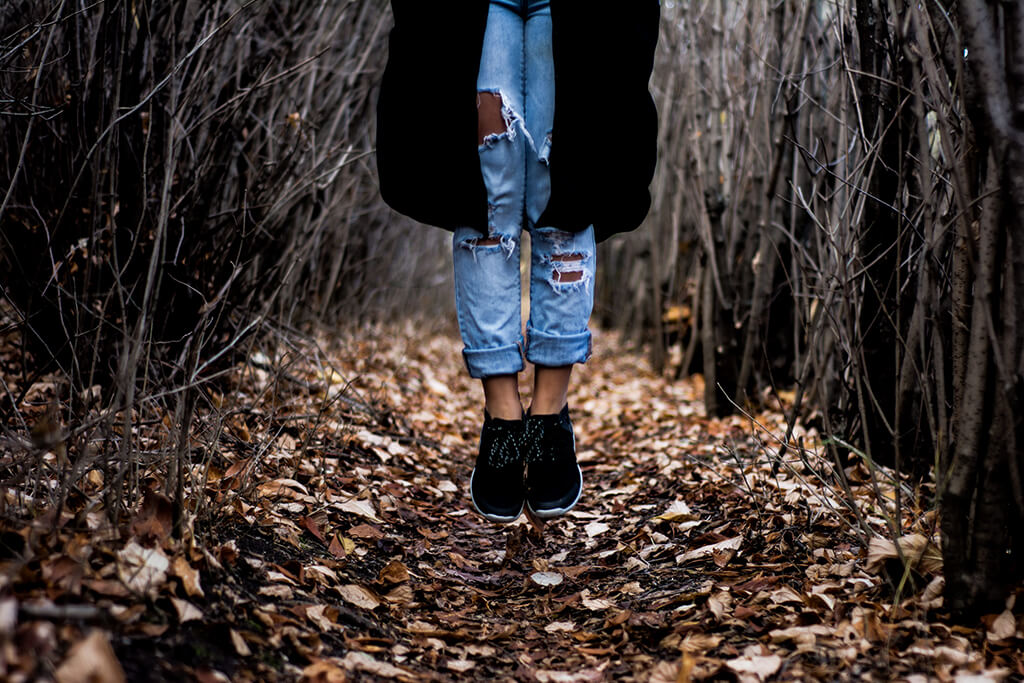 Someone in ripped jeans jumping on a leaf-covered woodland path