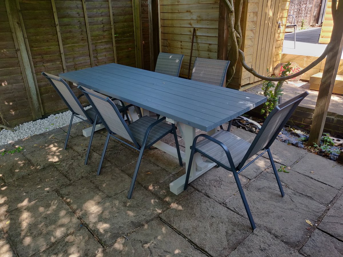 The grey-topped outdoor dining table with white trestle legs and six chairs on the patio, with the workshop behind