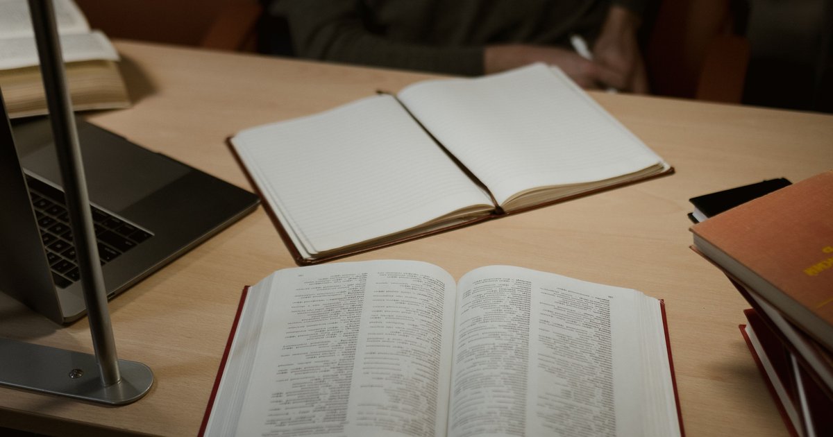 An open reference book and a blank notebook on a wooden desk beside a laptop and a stack of books