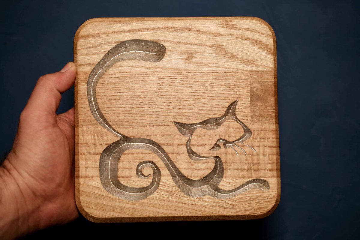 A square oak board carved with a flowing abstract cat outline, highlighted in silver, held up against a blue background