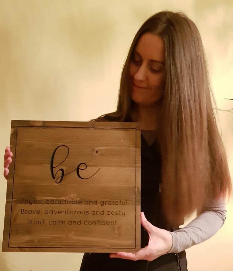 Caroline holding the finished display, a dark-stained pine board carved with the word be in script above three lines of text