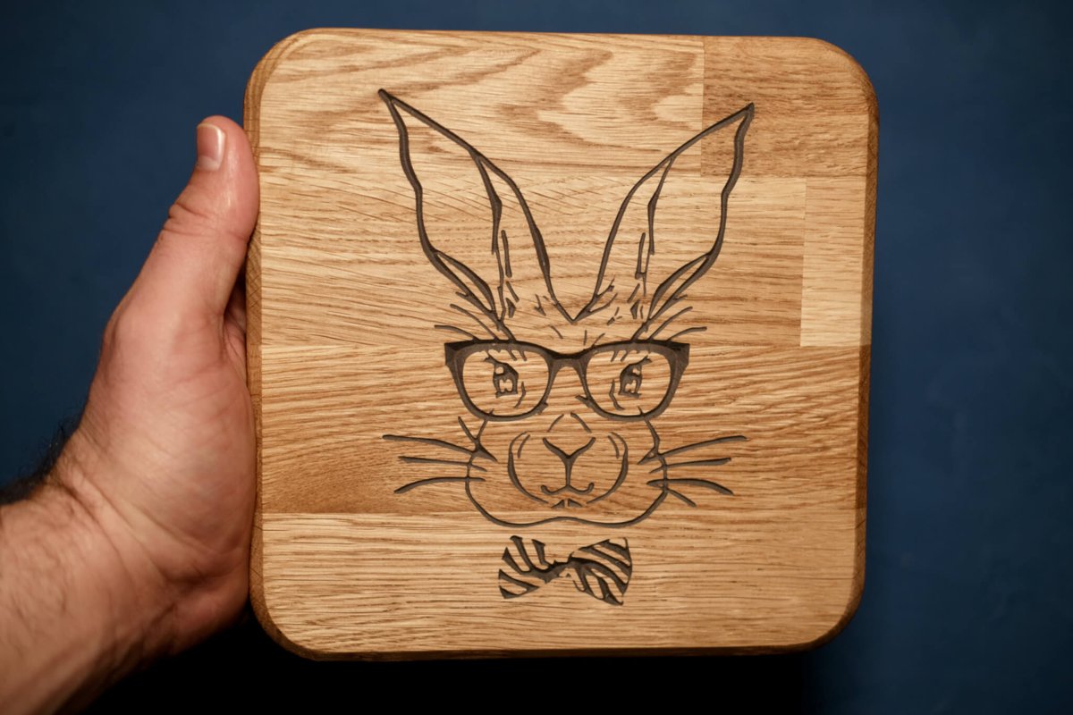 A square oak board carved with a rabbit wearing glasses and a bow tie, held up against a blue background