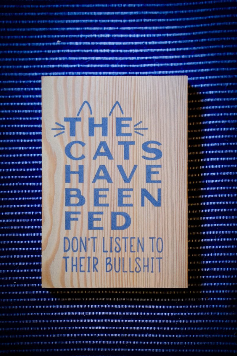 A pine board with The Cats Have Been Fed, Don't Listen To Their Bullshit painted in blue, with cat ears and whiskers on the first word