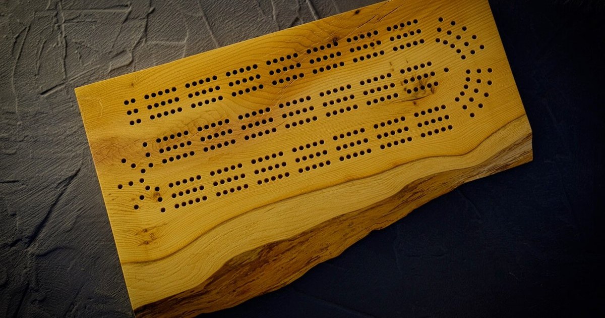 A live-edge yew cribbage board with rows of peg holes, photographed on a dark textured surface