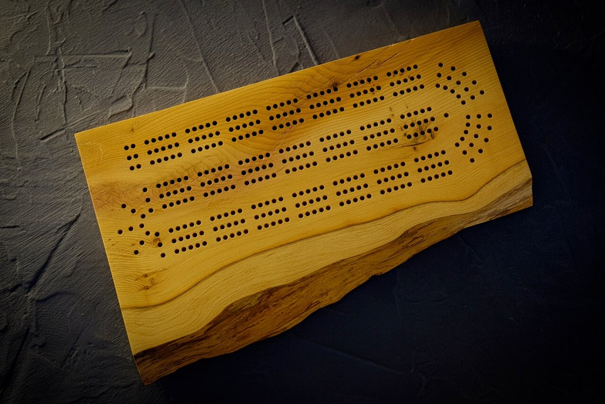 A live-edge yew cribbage board with rows of peg holes, photographed on a dark textured surface