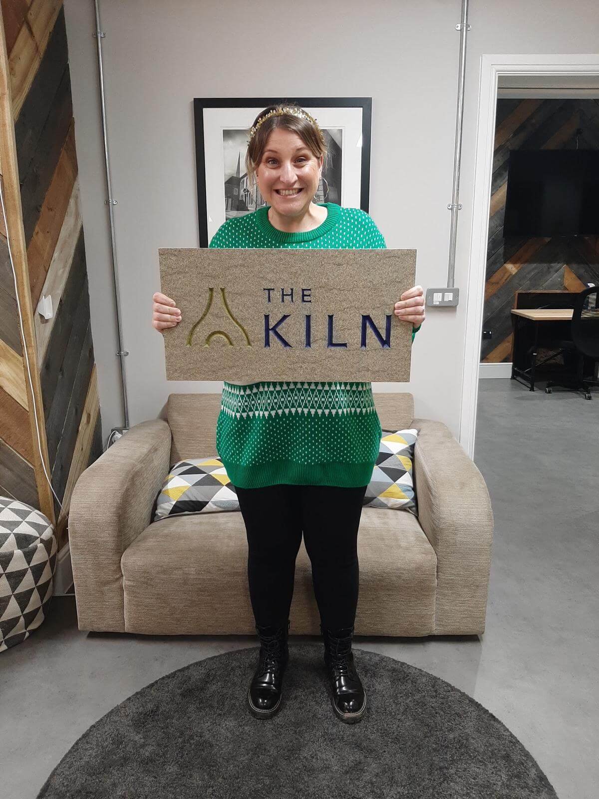 A woman in a green Christmas jumper and a gold crown holding the Kiln sign up in the co-working space's lounge