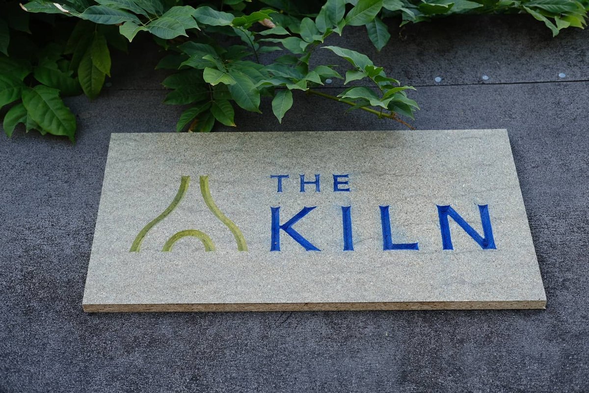 A chipboard sign carved with The Kiln's logo in green and the words The Kiln in blue, lying on a grey surface under a wisteria