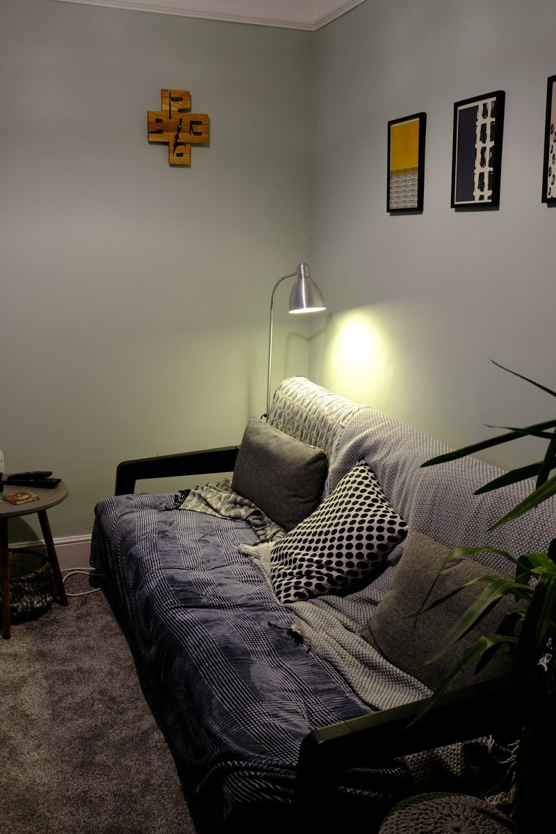 The clock on the wall above a sofa in the corner of the living room, beside a floor lamp and three framed prints