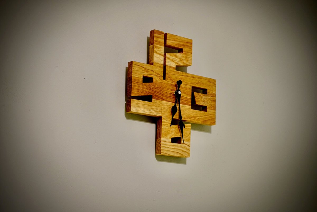 A wall clock made from interlocking blocks of oak with square cut-outs, with black hands, hanging on a grey wall