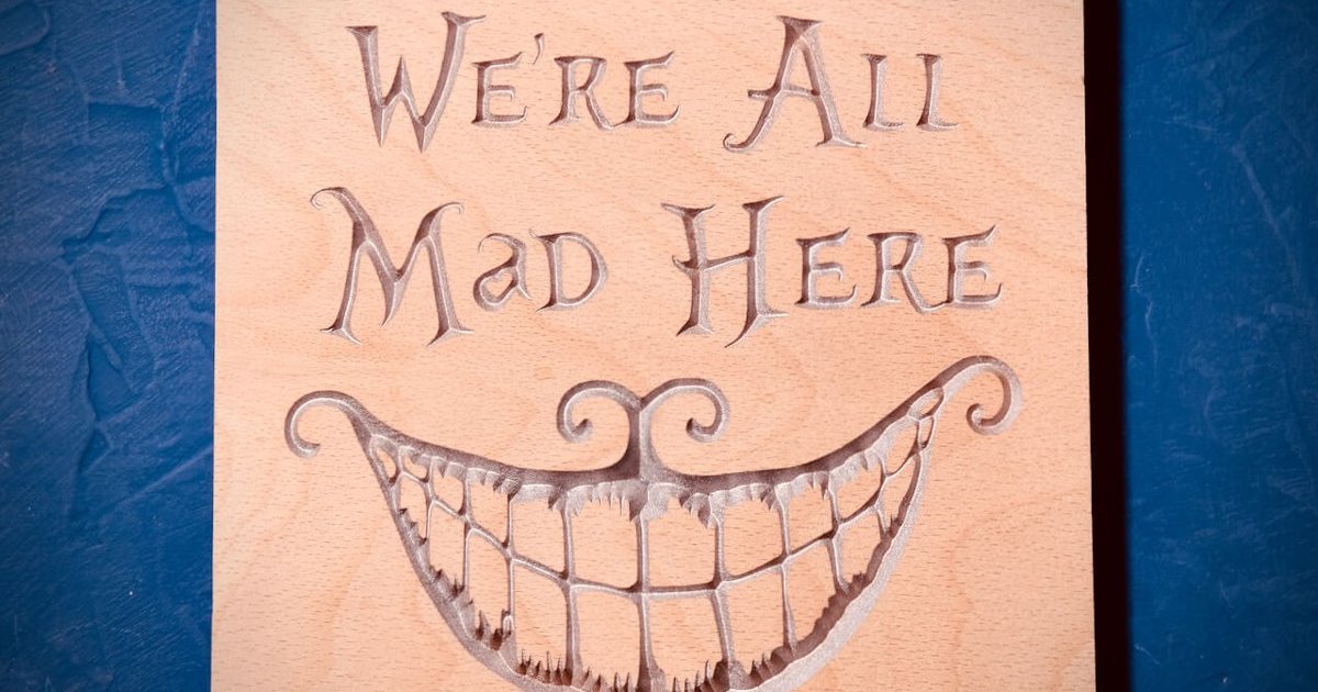 A beech board carved with We're All Mad Here and a wide Cheshire Cat grin, highlighted in silver, on a blue background