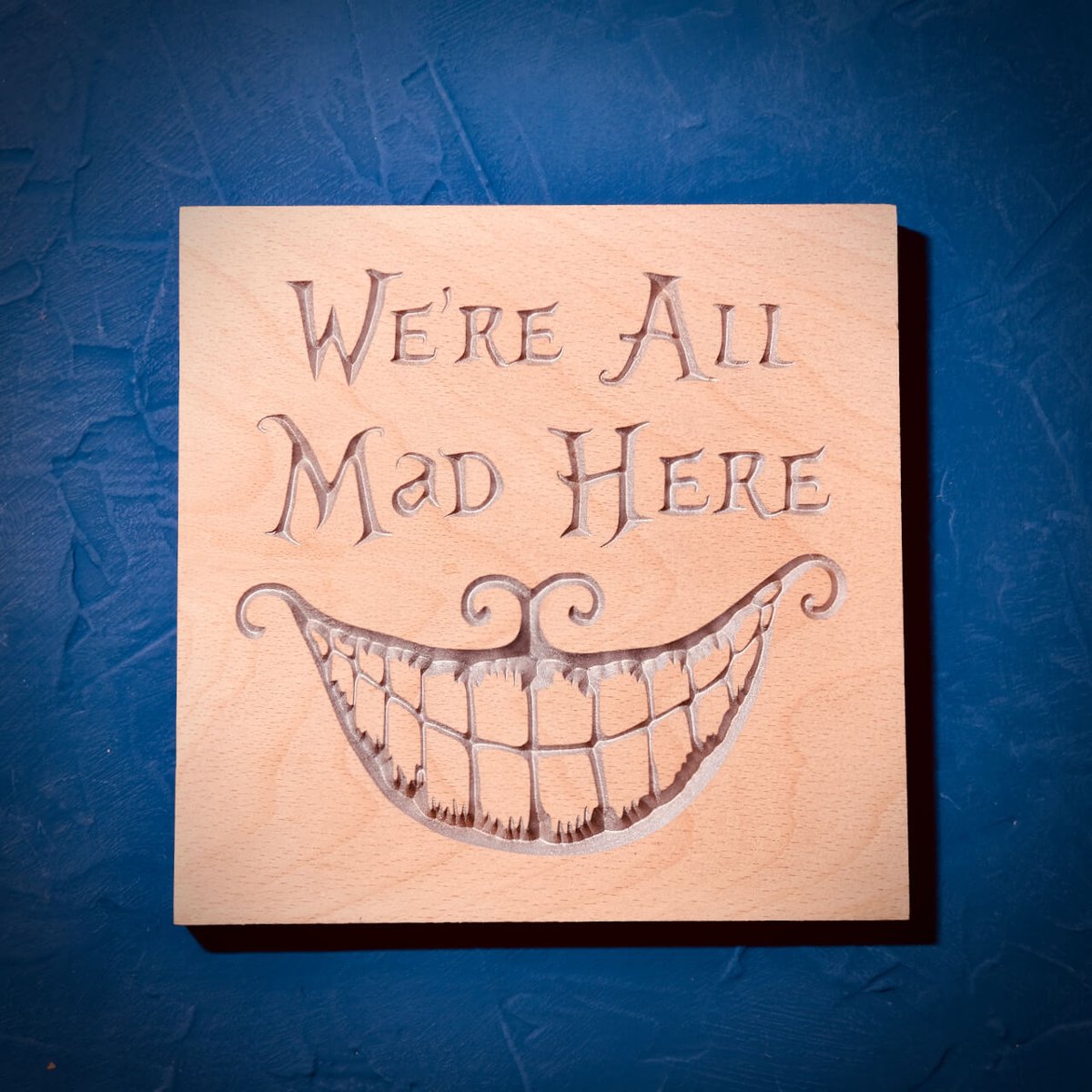 A beech board carved with We're All Mad Here and a wide Cheshire Cat grin, highlighted in silver, on a blue background
