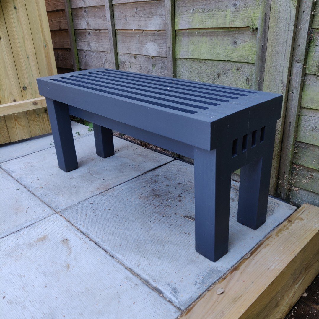 A slatted garden bench painted dark grey, standing on paving slabs in front of a fence
