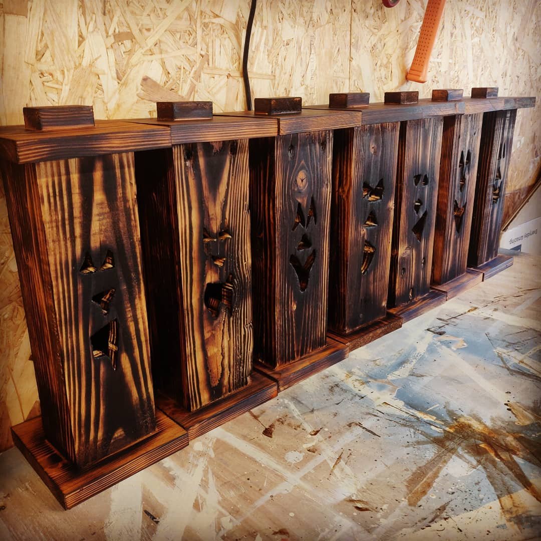 Seven tall jack-o'-lantern lanterns made from scorched pallet wood, with triangular eyes and jagged mouths cut into them, lined up on the workbench