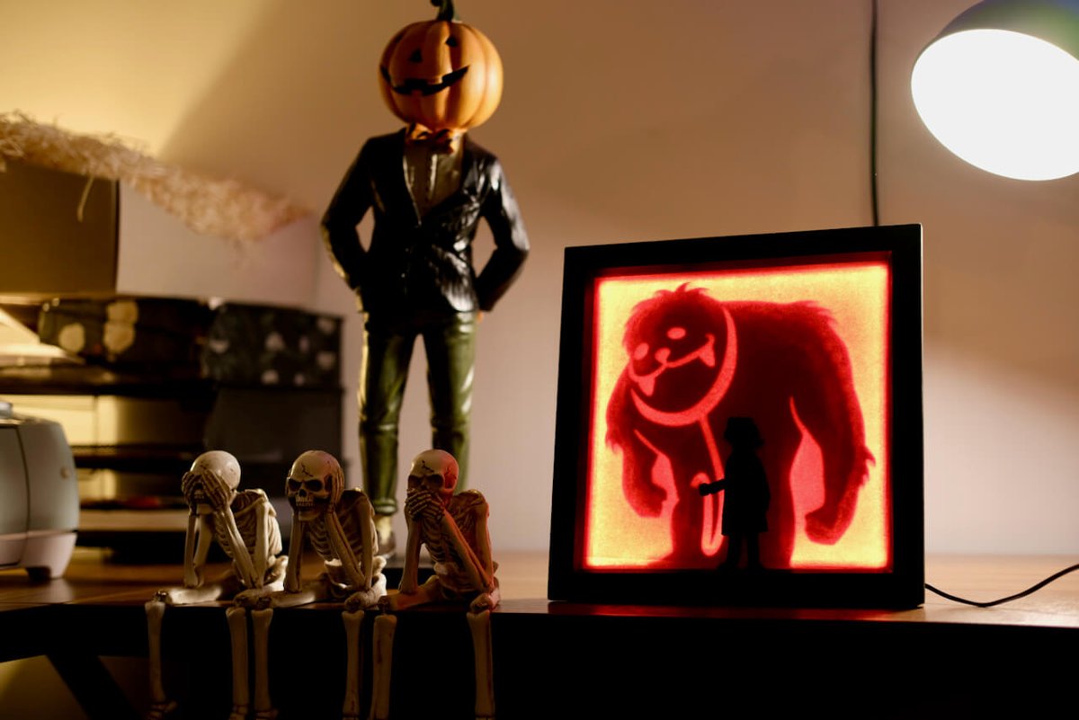 A light box glowing red with a monster silhouette behind a small girl, on a shelf beside skeleton figures and a pumpkin-headed statue