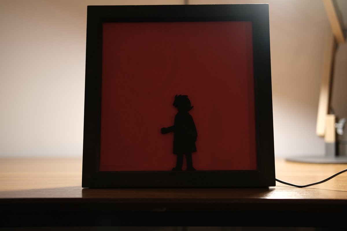 A black square frame with a red panel, showing only the silhouette of a small girl, on a desk with the light off