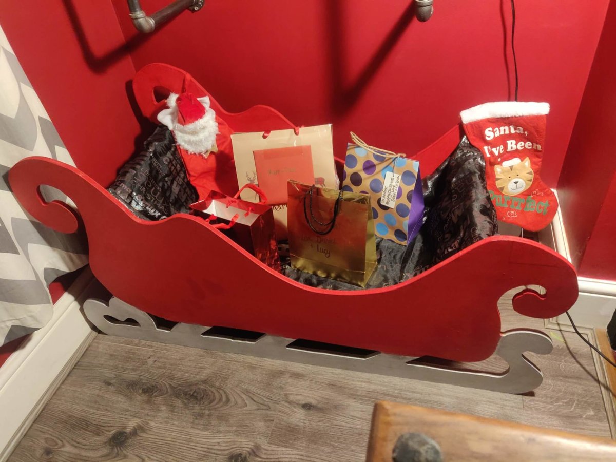 The finished sleigh, red with silver runners, full of wrapped presents and gift bags against a red wall