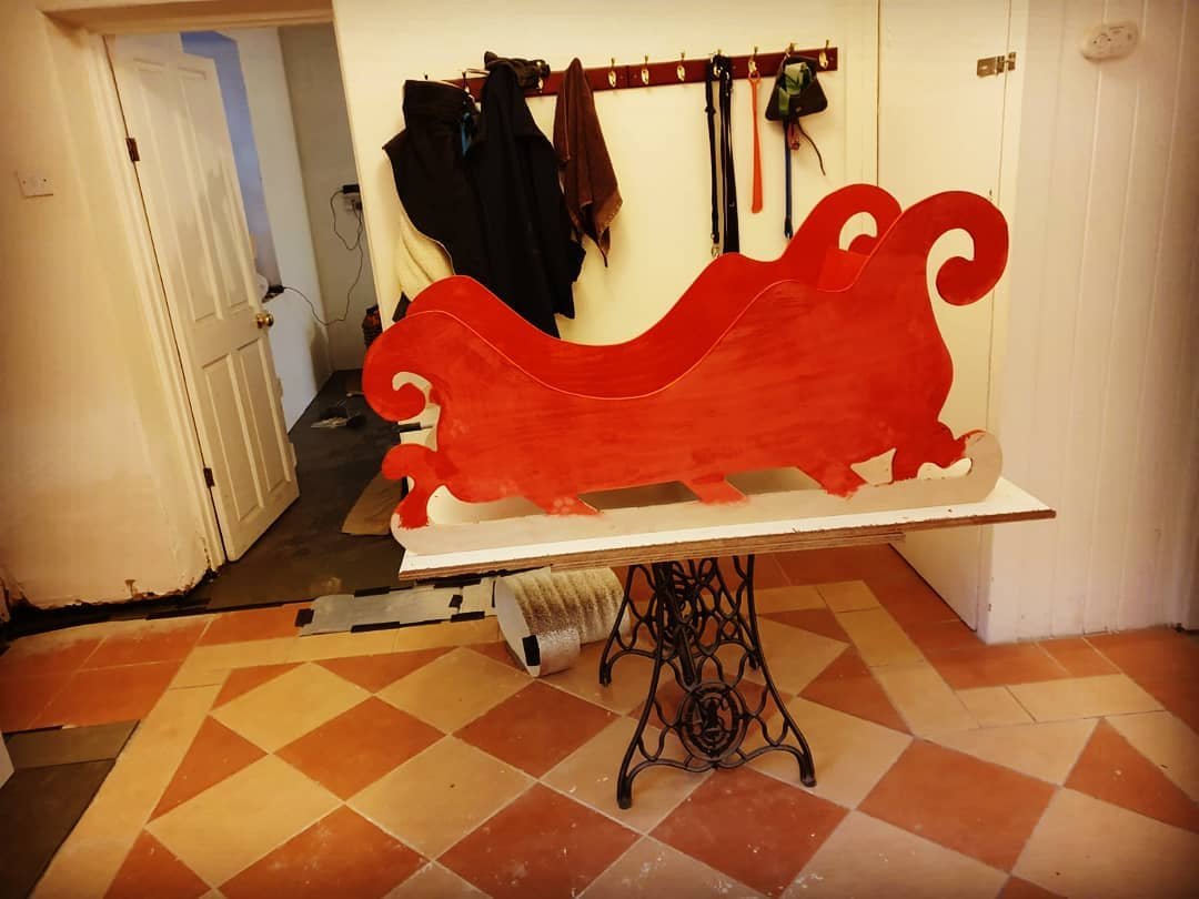 The sleigh's sides painted bright red, drying on a board balanced on a sewing machine stand in a tiled hallway