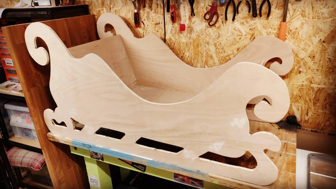 A plywood sleigh with curled runners and scrolled sides on a workbench in the workshop