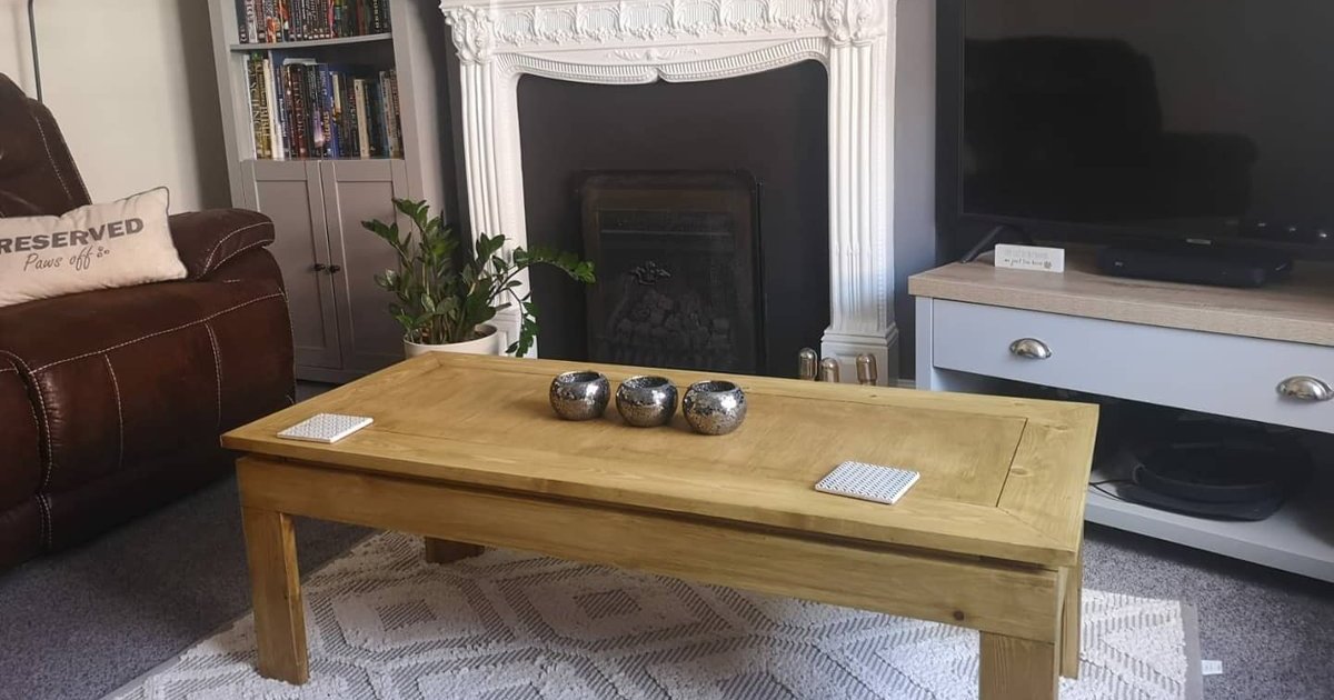 A pine coffee table with a framed top standing on a cream rug in front of a white fireplace