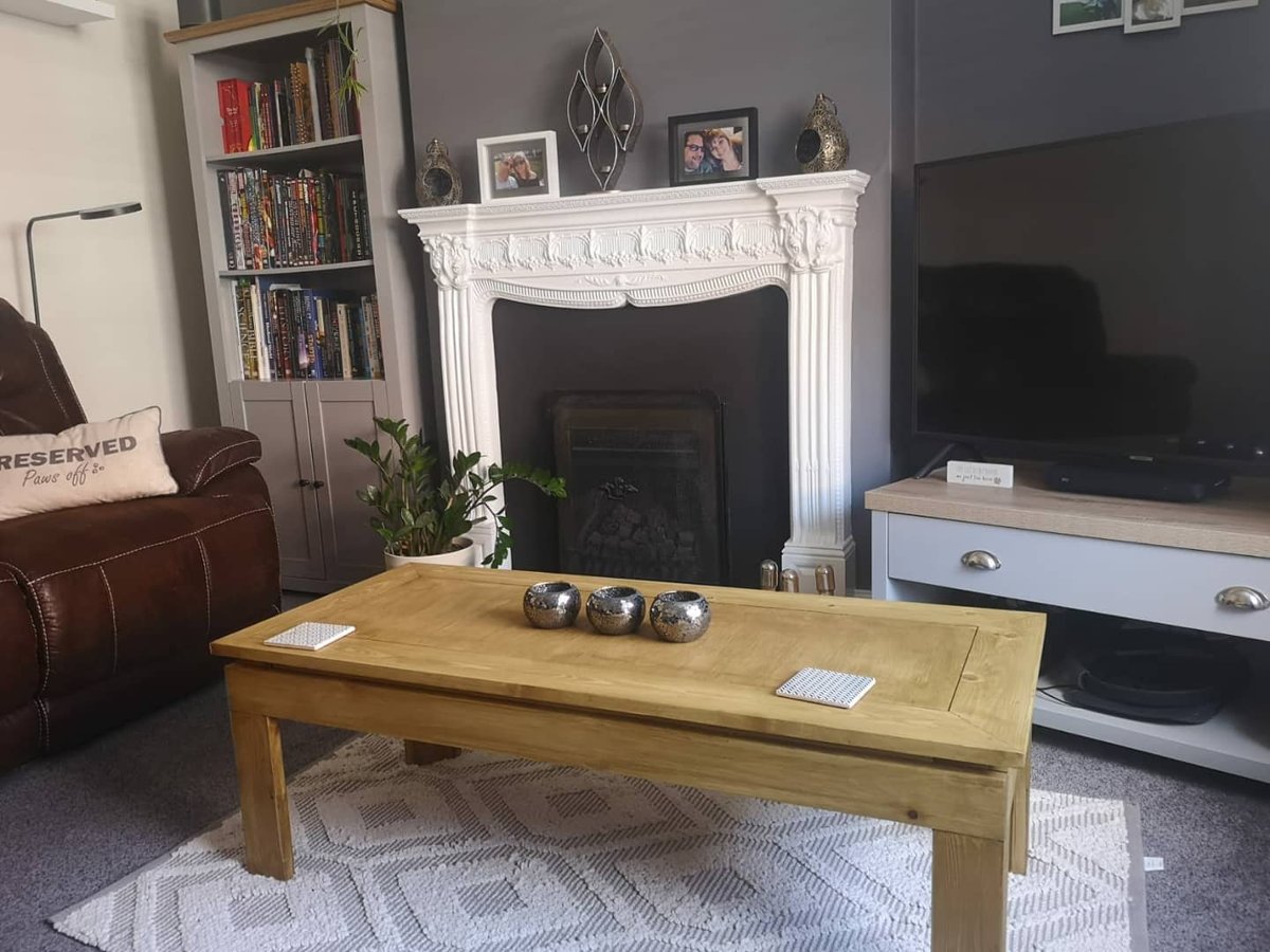 A pine coffee table with a framed top standing on a cream rug in front of a white fireplace