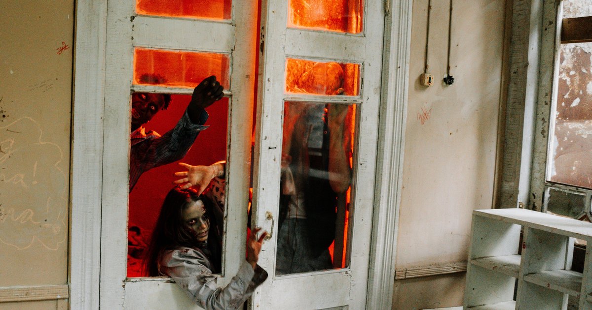 Zombies forcing their way through the broken panes of a pair of glass doors in a derelict house, lit red from behind